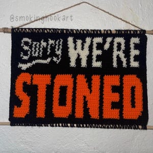 May include: Black and orange crocheted wall hanging with the text "Sorry WE'RE STONED" in white and orange. The sign is rectangular and hangs from a wooden dowel with a brown string. The background is a textured white wall.