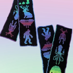 May include: Black crochet scarf with colorful alien and cryptid designs. The scarf features a variety of creatures, including an alien head, a bigfoot, and a deer. The designs are in shades of blue, purple, and green.