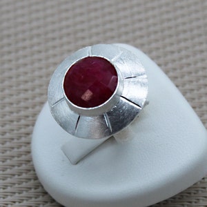 Peut inclure: Une bague en argent avec une pierre précieuse rouge sertie dans un design circulaire. La bague est sur un présentoir blanc.