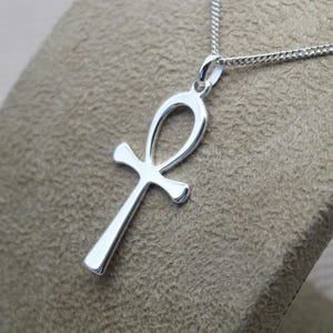 Può includere: Una collana con un ciondolo Ankh in argento. Il ciondolo è un design semplice e aperto con un anello in alto e una forma a croce in basso.