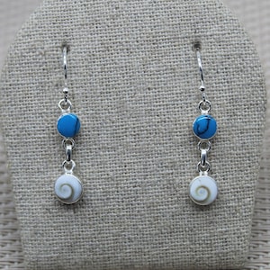 Peut inclure: Une paire de boucles d'oreilles pendantes en argent avec des accents de coquillages bleus et blancs. Les boucles d'oreilles présentent une petite perle ronde bleue et une plus grosse perle de coquillage blanche avec un motif en spirale.
