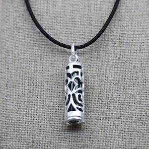 Può includere: Una collana con pendente color argento con un cordino nero. Il pendente è di forma cilindrica con un design traforato decorativo. Lo sfondo è un tessuto beige neutro.