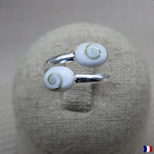 Pode incluir: Um anel prateado com dois cabochões brancos com padrão espiral. A banda do anel envolve o dedo, com os cabochões na parte superior. O anel é exibido numa superfície de tom neutro.