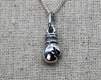 May include: A silver-colored boxing glove pendant hangs from a delicate silver chain. The pendant is a detailed miniature of a boxing glove, with visible stitching and a polished finish. The chain is a fine, linked design, suitable for everyday wear.