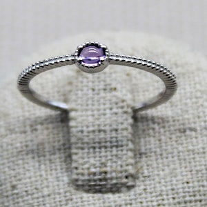 Peut inclure: Une bague en argent avec une petite pierre précieuse ronde de couleur violette sertie dans un sertissage à bélière. La bande de la bague a un design texturé.