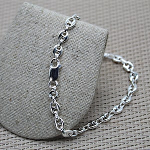 Könnte beinhalten: Ein silbernes Kettenarmband mit einem Ankerketten-Design. Das Armband liegt auf einem hellbraunen Stoffhintergrund.