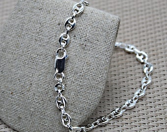 Puede incluir: Un brazalete plateado con un diseño de cadena de granos de café. El brazalete se muestra sobre una tela beige. El cierre es de tipo pinza rectangular. El metal brillante refleja la luz.