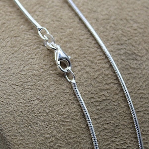 Peut inclure: Gros plan sur un collier chaîne serpent en argent avec un fermoir mousqueton. La chaîne est fine et souple, avec une surface lisse et réfléchissante. Le fermoir est également argenté et relie solidement la chaîne.