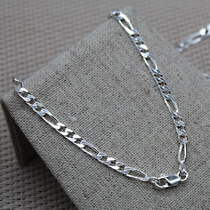 Puede incluir: Un collar de cadena de plata con un diseño clásico de eslabones figaro. La cadena se muestra sobre una superficie de tela texturizada.