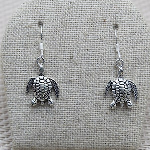 Può includere: Un paio di orecchini pendenti in argento con un designo di tartaruga dettagliato. Le tartarughe sono rivolte in avanti con le zampe allungate.