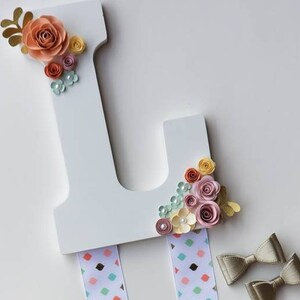 May include: White wooden letter L decorated with paper flowers in shades of pink, peach, and yellow. The letter has a ribbon attached to the bottom for hanging.