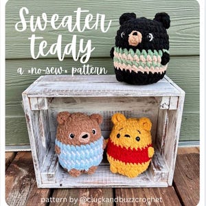 Può includere: Orsetti di peluche all'uncinetto in vari colori, tra cui marrone, nero e giallo, con dettagli di maglioni blu, verdi e rossi. Gli orsetti sono esposti in una cassa di legno bianca. Il testo sull'immagine recita "Sweater Teddy" e "a *no-sew* pattern."