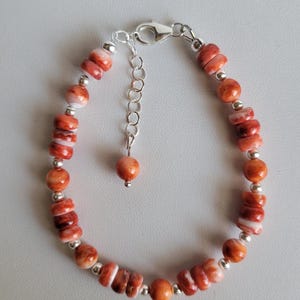 May include: A bracelet featuring round and disc-shaped beads in shades of orange and white with a marbled appearance. The beads are interspersed with small, polished silver-colored metal spheres. It includes a silver-colored lobster claw clasp.