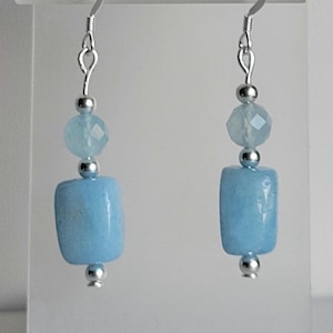 May include: A pair of dangle earrings featuring light blue, rectangular beads and faceted round beads. The earrings are suspended from silver-colored hooks and have small silver-colored accents. The earrings are displayed against a white background.