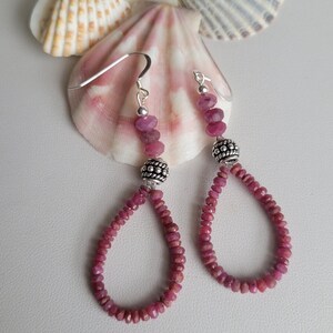 May include: A pair of dangle earrings featuring small, faceted, ruby-colored beads. The earrings have a teardrop shape and are accented with silver-colored metal beads and ear wires. The background includes seashells.