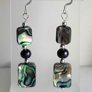 May include: Earrings with iridescent abalone shell squares and dark gray pearl beads. The earrings have silver-colored hooks and small silver-colored beads between the abalone and pearl elements. The earrings are displayed on a clear plastic stand.