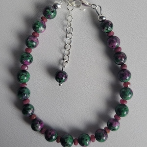May include: A beaded bracelet featuring round, mottled green and magenta zoisite stones interspersed with small, faceted dark pink beads. The bracelet includes a silver-toned metal lobster clasp and an adjustable chain with a single matching bead.