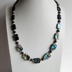 May include: A necklace displayed on a white bust. The necklace features rectangular abalone shell beads with iridescent colors, interspersed with black beads and silver-toned spacers. The necklace is a jewelry item.