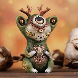May include: A whimsical green figurine with three eyes, small antlers, and gold accents. The creature holds a leaf and has a speckled design. The figurine is a unique art piece.