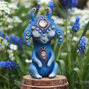 May include: A blue fantasy creature sculpture with multiple eyes and ear-like protrusions. The sculpture has a detailed design with white and gold accents. It is sitting on a wooden base, surrounded by flowers.