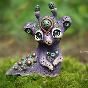 May include: A whimsical, handcrafted purple creature figurine with large green eyes and decorative details. The sculpture features a unique design with multiple eye-like features and a textured surface, set against a blurred green background.