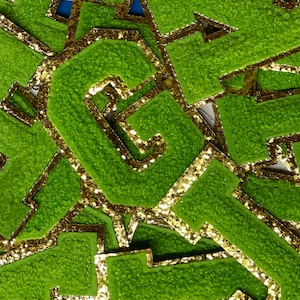 May include: A close-up of several green felt letter patches with gold glitter trim. The letters are stacked on top of each other, and the gold glitter trim is visible around the edges of each letter.
