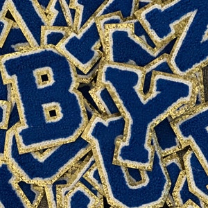 May include: A close-up of a pile of blue felt letters with gold glitter trim. The letters are in various sizes and are arranged in a random order. The letters are all uppercase and include the letters B, Y, and D.