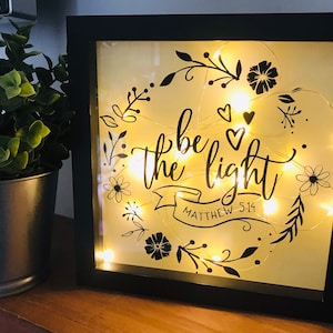 May include: A black framed light-up sign with a white background and black floral designs. The sign says "be the light" with a banner underneath that says "Matthew 5:14".