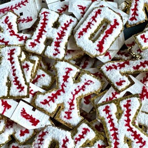 May include: A pile of white felt letters with red stitching and gold glitter. The letters are shaped like baseballs and are arranged in a random order.