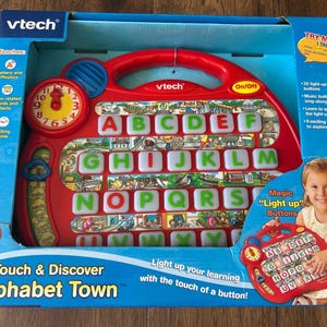May include: A red VTech Touch & Discover Alphabet Town educational toy in its original blue packaging. The device features a clock, twenty-six light-up letter buttons, and a town-themed illustration. The box includes text like Light up your learning.