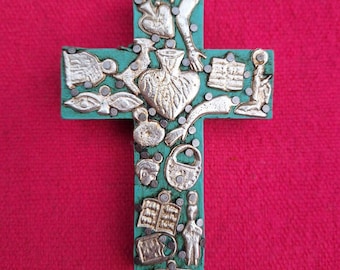 May include: A teal wooden cross decorated with silver metal charms. The charms include hearts, keys, a padlock, a bottle, and other religious symbols.