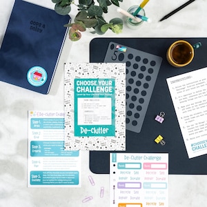 Puede incluir: Una composición de escritorio con un cuaderno azul, un diario titulado "Choose Your Challenge" y una plantilla gris. El diario tiene una cubierta turquesa y blanca con las palabras "De-clutter" y "Choose Your Challenge". También hay pegatinas y una taza de café.