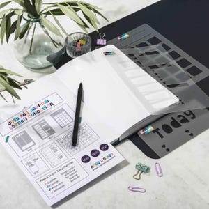 Può includere: Un diario aperto con una penna nera, stencil e fermagli. Il diario ha una guida alla collezione di stencil con il testo "Just Add Journal Stencil Collection". Gli stencil sono grigi e hanno varie forme e disegni. Un vaso di vetro con foglie verdi è sullo sfondo.