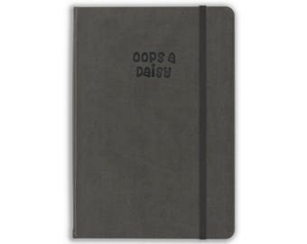May include: A dark gray notebook with a smooth, matte cover featuring the text "oops a daisy" embossed in the center. The notebook includes a vertical black elastic band closure attached to the right side, keeping the covers securely closed.