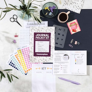 Può includere: Un layout piatto di un kit di progetto di diario con vari articoli di cancelleria. Il kit include un diario, adesivi, stencil, una penna e una tazza di caffè. Il diario recita "JOURNAL PROJECT KIT" e "Commonplace".