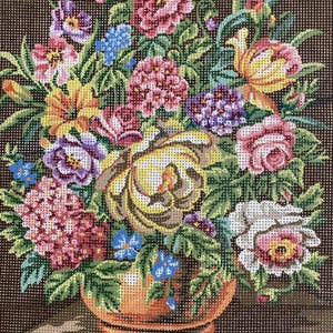 Puede incluir: Imagen de punto de cruz de un arreglo floral en un jarrón color cobre. El ramo presenta flores rosas, amarillas, moradas y azules, sobre un fondo marrón oscuro. El punto de cruz da una apariencia texturizada.