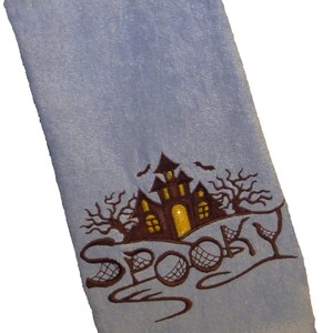 Könnte beinhalten: Ein hellblaues Handtuch mit einem gestickten Design eines Spukhauses mit einem gelben Fenster und dem Wort "Spooky" in brauner Kursivschrift.