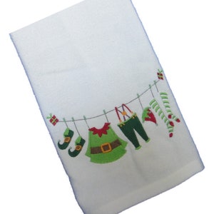 Könnte beinhalten: Weißes Weihnachtshandhandtuch mit gestickter Elfenkleidung, die an einer Wäscheleine hängt. Das Design umfasst grüne Elfenschuhe, eine grüne Tunika, grüne Hosen, einen roten Elfenhut und gestreifte Socken.
