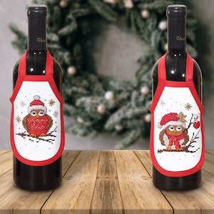 May include: Two red wine bottle covers with white fabric and embroidered images of owls wearing Santa hats. The owls are on branches with snowflakes and other wintery designs.