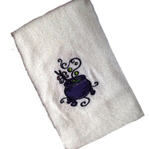 Könnte beinhalten: Ein weißes Küchentuch mit einem gestickten Design eines violetten Kessels mit grüner, sprudelnder Flüssigkeit und Dampf. Der Kessel hat einen schwarzen Griff und ist von schwarzen Spiralen umgeben.