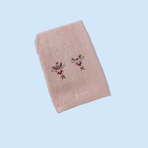 Könnte beinhalten: Ein rosa Handtuch mit zwei gestickten Rentiergesichtern. Die Rentiere haben braune Geweihe und rote Nasen.
