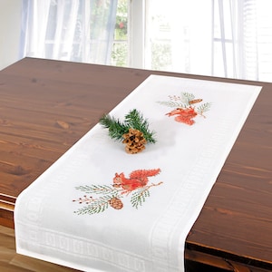 May include: White table runner with embroidered red squirrels on pine branches. The runner features a green and brown pine cone design.