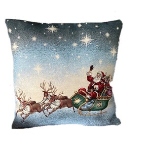 Könnte beinhalten: Ein quadratisches Zierkissen mit einer Weihnachtsszene. Der Weihnachtsmann winkt von seinem grünen Schlitten, der von Rentieren gezogen wird, vor einem sternenklaren blauen Himmel. Das Kissen hat ein strukturiertes Aussehen mit einem Farbverlauf von Blau und Weiß.