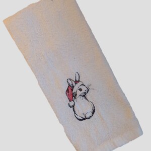 Könnte beinhalten: Cremefarbenes Handtuch mit einem gestickten Hasen, der einen roten Weihnachtsmannhut trägt. Der Hase ist mit schwarzem Faden umrandet, der Hut hat eine weiße Bommel. Das Handtuch hat eine strukturierte Oberfläche und einen gefalteten Rand.