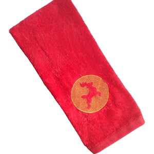 Könnte beinhalten: Rotes Handtuch mit einem goldenen gestickten Kreis, der ein stilisiertes Tiermotiv enthält.