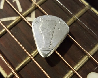 Könnte beinhalten: Ein silberner Gitarrenplektrum mit der Gravur "WARNER". Der Plektrum liegt auf den Saiten einer Gitarre.