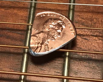 Könnte beinhalten: Ein Gitarrenplektrum aus Kupfer, hergestellt aus einem US-Cent, liegt auf den Saiten einer Akustikgitarre. Der Cent zeigt das Profil eines Mannes und die Worte "IN GOD WE TRUST". Das Plektrum hat einen silbernen Rand.