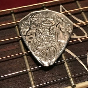 Könnte beinhalten: Ein silberner Gitarren-Plektrum in Form einer Münze mit einem Wappen-Design und der Nummer 50 darauf.