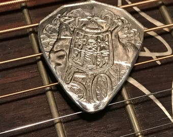 Könnte beinhalten: Ein silberner Gitarren-Plektrum in Form einer Münze mit einem Wappen-Design und der Nummer 50 darauf.