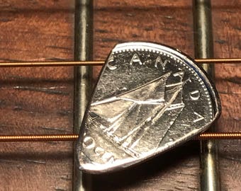 Könnte beinhalten: Ein Gitarrenplektrum aus einer kanadischen Münze, das auf den Saiten und dem Griffbrett einer Akustikgitarre liegt. Die Münze zeigt ein Segelschiff und das Wort "CANADA". Das Plektrum ist silberfarben.